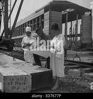 Slaviansk, Donetsk region, Ukrainian SSR, Soviet Union. Late 1960s. An engineer and two female workers in white coats discussing blueprints at the construction site of the AIZ factory glass workshop. They stand near a gantry crane, using a concrete block as a table. The block bears a production date marking "17.XI.66". In the background, the concrete skeleton of the unfinished industrial building is visible. This archival black and white photograph captures the teamwork and industrial development in the peaceful Donbass region long before the war Stock Photo