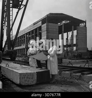 Slaviansk, Donetsk region, Ukrainian SSR, Soviet Union. Late 1960s. An engineer and two female workers in white coats discussing blueprints at the construction site of the AIZ factory glass workshop. They stand near a gantry crane, using a concrete block as a table. The block bears a production date marking "17.XI.66". In the background, the concrete skeleton of the unfinished industrial building is visible. This archival black and white photograph captures the teamwork and industrial development in the peaceful Donbass region long before the war Stock Photo