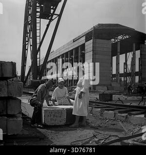 Slaviansk, Donetsk region, Ukrainian SSR, Soviet Union. Late 1960s. An engineer and two female workers in white coats discussing blueprints at the construction site of the AIZ factory glass workshop. They stand near a gantry crane, using a concrete block as a table. The block bears a production date marking "17.XI.66". In the background, the concrete skeleton of the unfinished industrial building is visible. This archival black and white photograph captures the teamwork and industrial development in the peaceful Donbass region long before the war Stock Photo