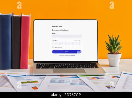 Open laptop displaying login screen on desk with documents, books, and plant, on orange backdrop Stock Photo