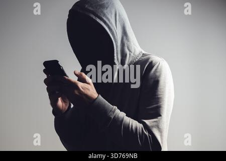 Man in hoodie using smartphone in dark shadow, face hidden in black, concept of anonymity, secrecy, cybercrime, or privacy. Selective focus. Stock Photo