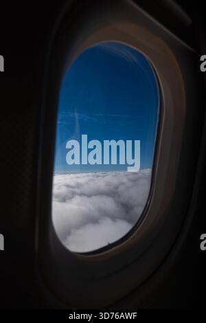 Vertical shot of a clear blue sky above a dense layer of white clouds, framed perfectly by the dark, oval window of an airplane cabin, symbolizing tra Stock Photo