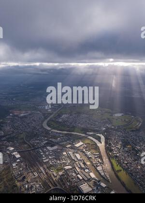 Dramatic aerial view of an industrial city and a winding river, with powerful crepuscular sun rays (God rays) breaking through dark storm clouds onto Stock Photo