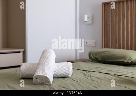 Three rolled towels arranged on a green sheet bed with wood headboard in modern bedroom. Stock Photo