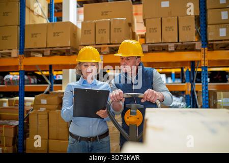 Older manager talking with worker in a warehouse, reading plan document. Stock Photo