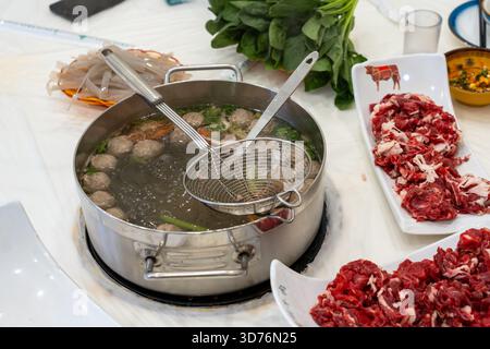 A stainless steel hot pot of simmering broth with meatballs sits on a table. Plates of thinly sliced raw beef and fresh vegetables await cooking, high Stock Photo