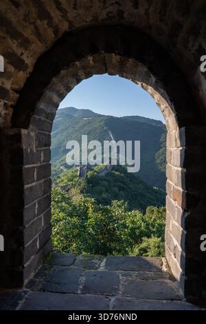 A dark stone arch window on the Great Wall frames a bright view of the winding wall and lush green mountains outside. The contrast emphasizes the hist Stock Photo