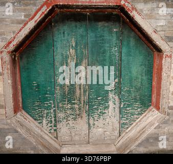 An octagonal window set in a brick wall is closed with wooden shutters that show heavily peeling green paint. The red and gray frame highlights the de Stock Photo