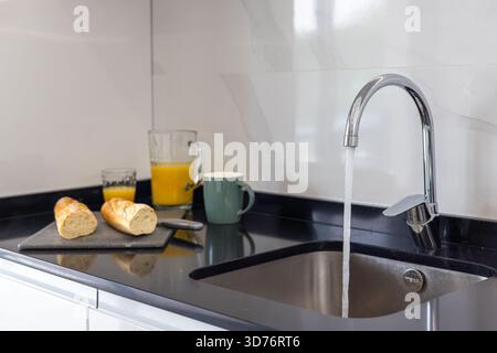 Modern kitchen sink with flowing tap water, orange juice, baguette, and ceramic mug on counter Stock Photo