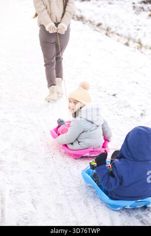 A woman pulls two children on brightly colored sleds through a snowy landscape. The children are bundled up warmly for a fun day outside enjoying the Stock Photo