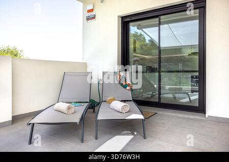 Minimalist outdoor seating area with loungers, towels, and reflection of kitchen in glass door Stock Photo