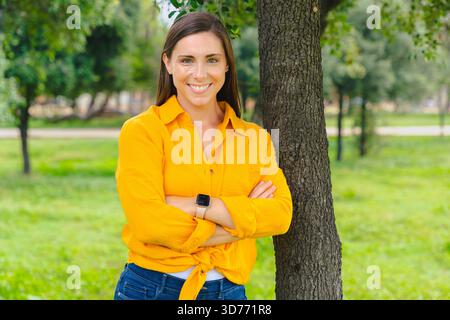 Smiling woman standing with arms crossed outdoors in park looking at camera Stock Photo