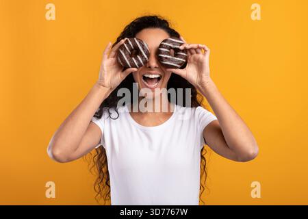 Happy Latin Woman Covers Eyes With Donuts in Bright Studio Setting Enjoying Sweet Treats Stock Photo