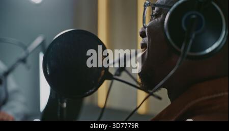 Close Up of African American Man in Glasses and Headphones Speaking Emotionally Into Professional Studio Microphone. Adult Man Smiles and Reacts Vividly While Recording Engaging Podcast Conversation. Stock Photo