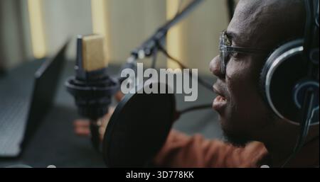 Close Up African American Man Wearing Glasses and Studio Headphones Speaking Energetically Into Pro Microphone. Laptop Sits on Table While Man Reacts With Expressive Emotion During Podcast Recording. Stock Photo