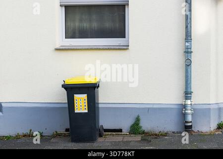 A black garbage bin with a yellow lid is positioned next to a plain building wall. A small section of grass is visible near the base, and the scene is Stock Photo
