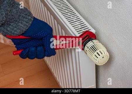 A person wearing blue gloves uses a wrench to adjust a heating radiator valve. This activity takes place in a cozy indoor setting, highlighting winter Stock Photo