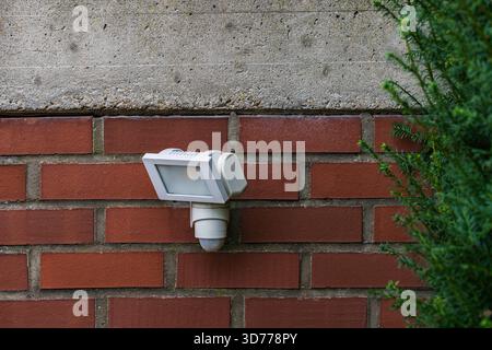A motion sensor light is mounted on a brick wall, providing security and illumination. The light is positioned next to lush green foliage, enhancing t Stock Photo