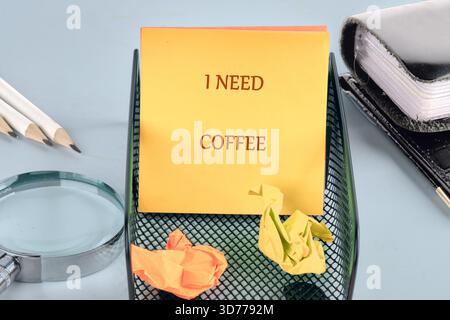 A bright yellow sticky note with I NEED COFFEE rests in a desk organizer beside crumpled papers, pencils, a magnifier, and a notebook on a soft blue w Stock Photo