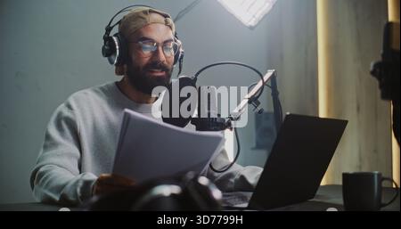 Professional Male Podcast Host Recording Episode in Studio. Bearded Man Wearing Glasses and Headphones Reads From Script Into Microphone in Audio Studio, Focusing on Production Digital Media Content. Stock Photo