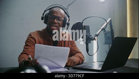 African American Male Expert Content Creator in Studio Records Podcast, Using Prepared Notes and Professional Audio Gear. Concept of Knowledge Sharing and Production of Expert Driven Digital Media. Stock Photo