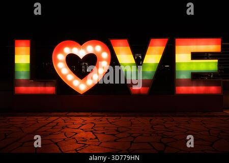 A colorful illuminated LOVE installation shines brightly in the dark, featuring a rainbow gradient and glowing heart-shaped letter. Stock Photo