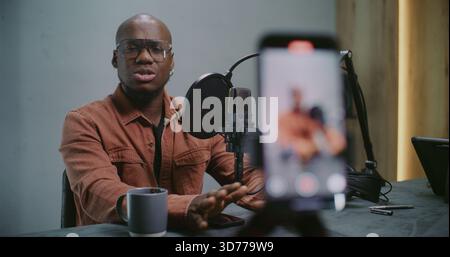 Process of Shooting Vertical Phone Footage in Which African American Man Speaking Confidently Into Mic While Gesturing Over Table With Cup and Smartphone. Concept of Live Podcast Moment. Rack Focus. Stock Photo