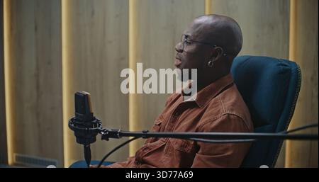 Close Up of Bald, Black Expert Speaker With Glasses and Earring, Positioned Professional Microphone in Modern Audio Studio. Concept of Professionalism of Digital Broadcasting and Thought Leadership. Stock Photo