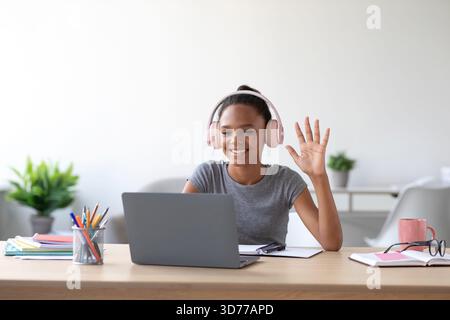 Smiling Student Studies at Home During Online Class While Waving at Laptop Camera Stock Photo