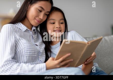 Happy Asian Girl Enjoys Reading With Mother on Cozy Sofa in Home Interior Stock Photo