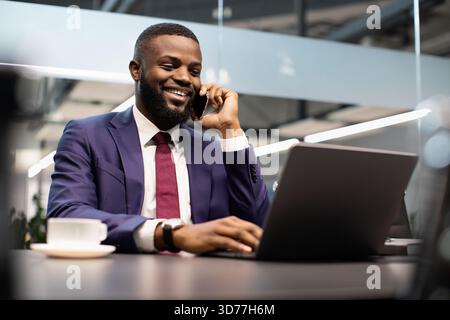 Smiling black manager working on laptop and talking on phone Stock Photo