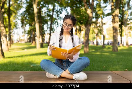 Young Woman Studying in a Park on a Sunny Day While Sitting on a Wooden Platform Surrounded by Green Trees Stock Photo
