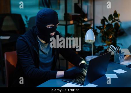 Young Adult Man Wearing Mask Stealing Data Using Laptop in Office Stock Photo