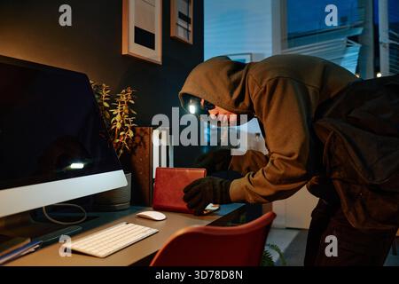 Young Adult Caucasian Man Stealing Wallet from Office Desk during Nighttime Stock Photo
