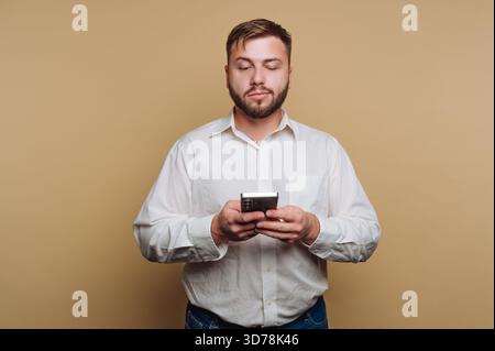 portrait of young man in a white shirt looks down at his smartphone in a relaxed manner on beige background Stock Photo