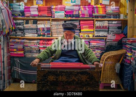 Lijiang, China - 21 November 2025: View of a woman weaving vibrant textiles in a cozy shop, surrounded by stacks of colorful fabrics and intricate pat Stock Photo