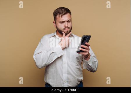 portrait of young man thoughtfully examines his smartphone while stroking his chin on beige background Stock Photo