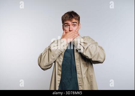 A young man stands with wide eyes and an open mouth, covering it with his hands, showing shock or disbelief in a neutral setting. Stock Photo
