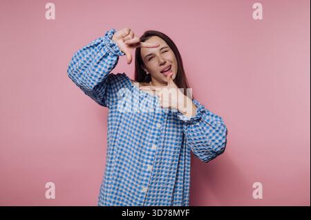 A young woman stands in front of a pink background, wearing a blue checkered dress. She strikes a playful pose, using her hands to mimic a camera fram Stock Photo