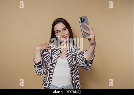 A young woman stands in front of a plain backdrop while taking a selfie. She wears a trendy jacket and denim jeans, smiling confidently as she capture Stock Photo