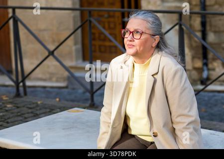 In a quaint, historic area, a woman with silver hair and glasses sits on a stone bench, wearing a light sweater and blazer. She gazes thoughtfully, en Stock Photo