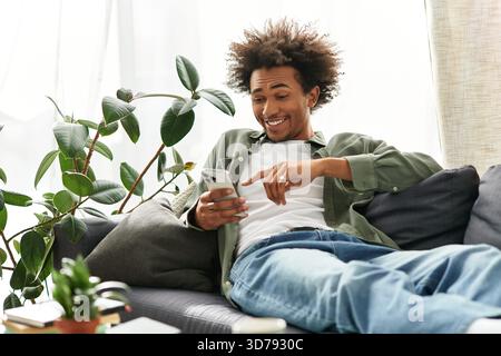 A young strong man laughs on his couch, using his smartphone in a stylish home. Stock Photo