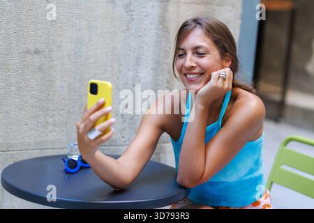 Young woman sitting alone at a cafe table outdoors, taking a selfie Stock Photo