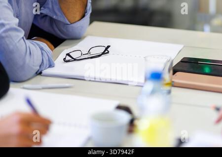 Participants collaborate during a business meeting with tools and materials on the table, showcasing glasses and stationery in an office setting Stock Photo