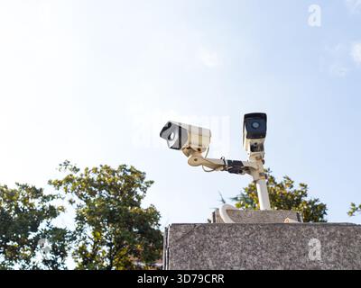 Two surveillance cameras mounted on a stone pillar, trees behind. Stock Photo