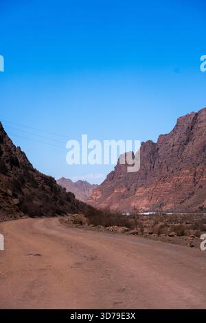 An unpaved road winding through a tight mountain pass or valley framed by massive, rugged red rock cliffs under a bright blue sky Stock Photo