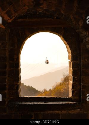 View of a cable car suspended in the distance, framed by an ancient stone archway and rolling hills bathed in soft light, Beijing, Beijing, China. Stock Photo