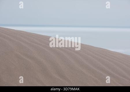 a sand dune with soft undulating patterns on the sand. In the background, you can see a calm sea or a large lake under a pale, cloudy sky. Stock Photo