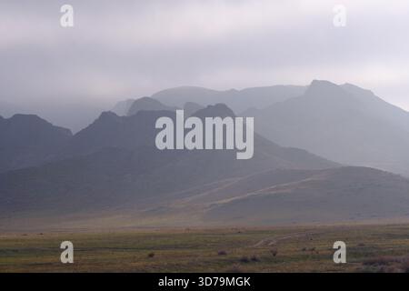 Gloomy mountain landscape, shrouded in fog. In the foreground is a field with a path, and the mountains in the distance are lost in the haze. The mute Stock Photo