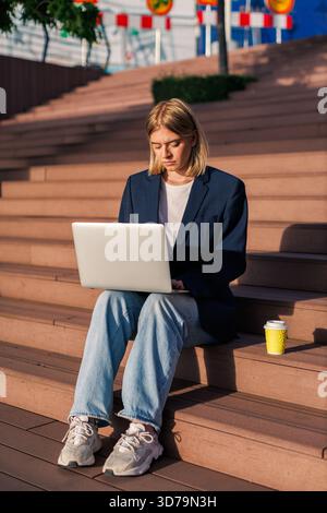 Young woman freelancer works on laptop while sitting on outdoor steps in a vibrant urban setting Stock Photo